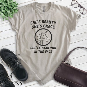 Puede incluir: Camiseta gris claro con el texto "She's Beauty She's Grace She'll Stab You In The Face" y un gr&aacute;fico de unicornio negro. La camiseta est&aacute; sobre una superficie de madera blanca, con zapatillas negras, un bolso marr&oacute;n y gafas.
