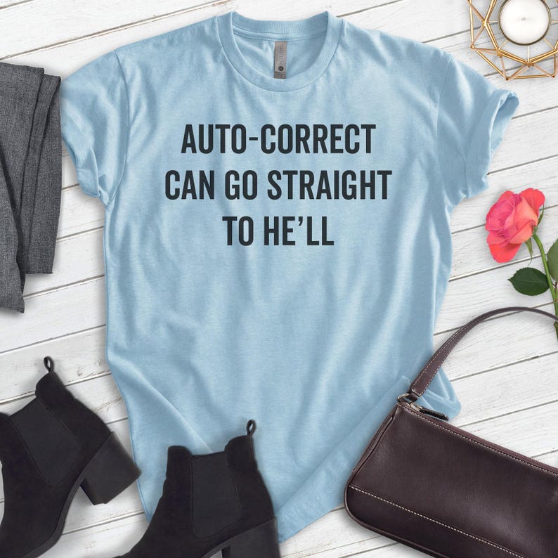May include: A light blue t-shirt with the text "AUTO-CORRECT CAN GO STRAIGHT TO HE'LL" in black. The shirt is displayed with black ankle boots, a brown purse, and a rose.