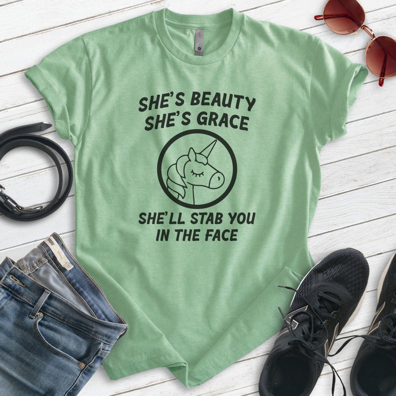 Puede incluir: Una camiseta verde claro con el texto "She's Beauty She's Grace She'll Stab You In The Face" y un gr&aacute;fico de unicornio negro. La camiseta se combina con vaqueros azules, un cintur&oacute;n negro, gafas de sol y zapatillas negras.
