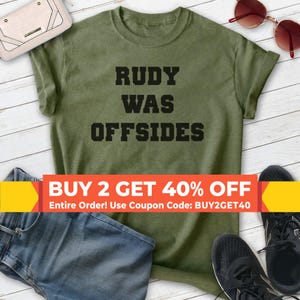 May include: A green t-shirt with the text "RUDY WAS OFFSIDES" printed in black. The shirt is photographed on a wooden surface with jeans and sneakers. The text "BUY 2 GET 40% OFF" is displayed in a yellow and red banner with the text "Entire Order! Use Coupon Code: BUY2GET40" below.