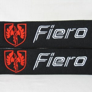 May include: Two black seatbelt pads with white embroidered "Fiero" text and a red embroidered logo.
