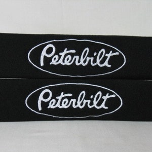 Op de afbeelding: Twee zwarte stoffen veiligheidsgordelpads met witte geborduurde ovale logo's met de tekst "Peterbilt".