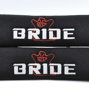 May include: Two black seat belt covers with the word "BRIDE" embroidered in white, and a red graphic above the text. The covers are made of a soft, textured material.