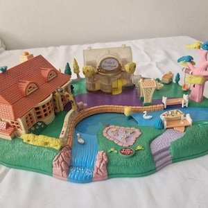Op de afbeelding: Een roze en witte plastic speelset met een huis, een boom, een vijver en een brug. De speelset is ontworpen voor een poppenhuis en heeft een klein huis met een rood dak, een boom met een roze stam en groene bladeren, een blauwe vijver met een kleine brug en een klein wit bootje. De speelset is gemaakt van plastic en is in goede staat.