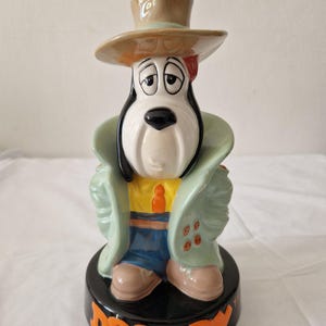 Puede incluir: Una figurita de cerámica de Droopy Dog, un personaje de dibujos animados, que lleva un abrigo verde, pantalones azules, un sombrero marrón y una corbata amarilla. La figurita está de pie sobre una base negra con la palabra "Droopy" escrita en letras naranjas.