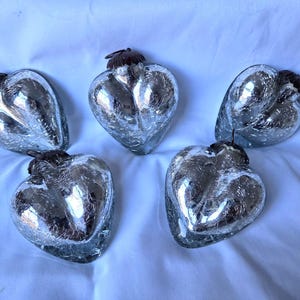 Puede incluir: Cinco adornos plateados en forma de corazón con efecto de vidrio craquelado. Cada adorno tiene una tapa marrón oscuro y un lazo negro para colgar. Los corazones están dispuestos sobre una superficie blanca.