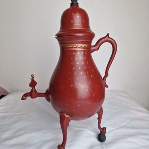 May include: A red metal teapot with a gold trim and a black knob on the lid. The teapot has a spout and a handle and sits on three legs.