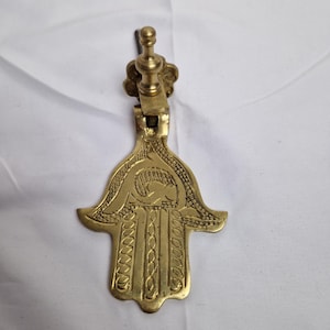 Peut inclure: Un heurtoir de porte en métal doré en forme de main de hamsa avec des détails complexes. Le hamsa est un symbole de protection et de bonne chance dans de nombreuses cultures.