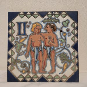 Puede incluir: Azulejo cerámico cuadrado con la representación de dos figuras, posiblemente Géminis, con el número romano II en azul. Las figuras están rodeadas de patrones florales y geométricos en verde, amarillo y azul, sobre un fondo blanco con borde azul oscuro.