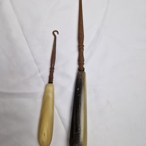 Puede incluir: Dos cuernos para zapatos antiguos hechos de cuerno y metal. El cuerno más grande es marrón oscuro y negro, mientras que el cuerno más pequeño es marrón claro. Ambos tienen un gancho de metal en la parte superior.