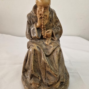 Puede incluir: Una figurita de cerámica de un monje sentado con una larga barba, vistiendo una túnica marrón y sosteniendo un rosario en sus manos.