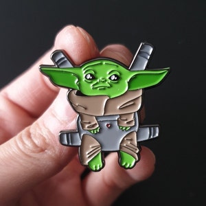 Puede incluir: Un pin de esmalte verde y beige que representa a Grogu, también conocido como Baby Yoda, de la franquicia Star Wars. El pin muestra a Grogu en su hover-pram, con una combinación de colores gris y beige. El pin está en la mano de una persona.