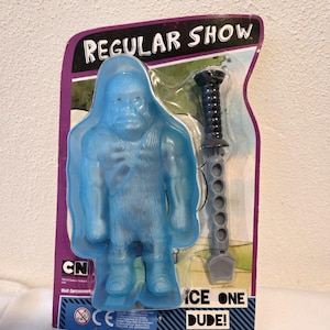 Puede incluir: Una figura de acción translúcida azul de un personaje parecido a un yeti de la serie "Regular Show", empaquetada con un arma gris. El embalaje presenta el logotipo del programa y el texto "ICE ONE DUDE!"