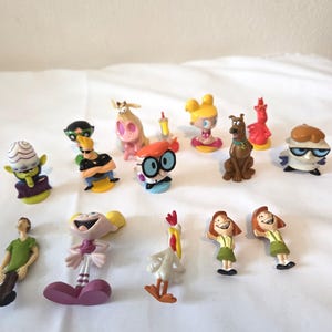 May include: A selection of colourful cartoon character figurines, including Dexter, Dee Dee, and Scooby-Doo. The figures are made of plastic and are set against a white background. The collection includes characters from various animated series.