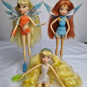 Puede incluir: Tres muñecas de hadas con alas y pelo largo. Las muñecas llevan atuendos coloridos y tienen diferentes colores de pelo. Las muñecas están en diferentes poses.