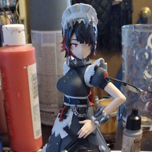 Op de afbeelding: Gedetailleerd beeldje van een personage in een dienstmeisjesoutfit, met zwarte en witte accenten, rood haar en een bijpassende basis. Accessoires zijn onder meer een zwart-wit mes en een zwart wapen met oranje details. Verfpotten op de achtergrond.