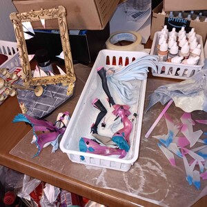 Puede incluir: Un espacio de trabajo desordenado con varios objetos. Un espejo dorado ornamentado roto, figuras de anime desmontadas en un contenedor blanco, botes de pintura y cajas de cart&oacute;n son visibles. Las figuras son principalmente rosas, azules y negras.