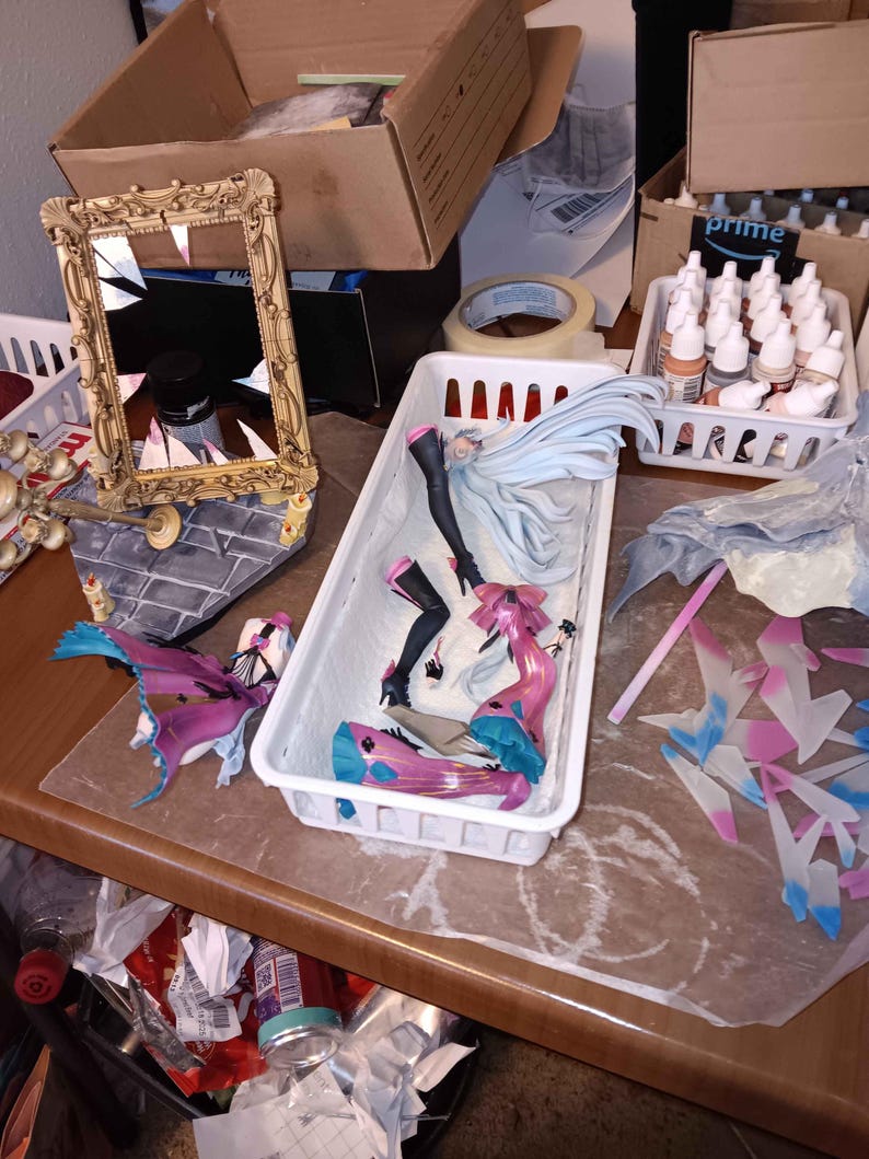 Puede incluir: Un espacio de trabajo desordenado con varios objetos. Un espejo dorado ornamentado roto, figuras de anime desmontadas en un contenedor blanco, botes de pintura y cajas de cart&oacute;n son visibles. Las figuras son principalmente rosas, azules y negras.