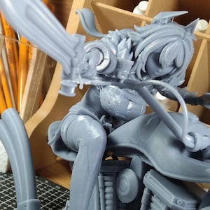 Op de afbeelding: Een grijze, onbeschilderde figuur van een vrouwelijk personage in een dynamische pose, mogelijk uit een videogame of anime. De figuur is gedetailleerd, met ingewikkelde bepantsering en een gestileerd ontwerp. De figuur zit, met één been gebogen en het andere uitgestrekt.