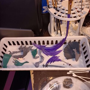 Puede incluir: Una bandeja de pl&aacute;stico blanco contiene varias figuras desmontadas y material de pintura. Las figuras son en tonos verdes, grises y morados. Los pinceles est&aacute;n en un soporte blanco. Una paleta de pintura con pintura gris est&aacute; en primer plano.