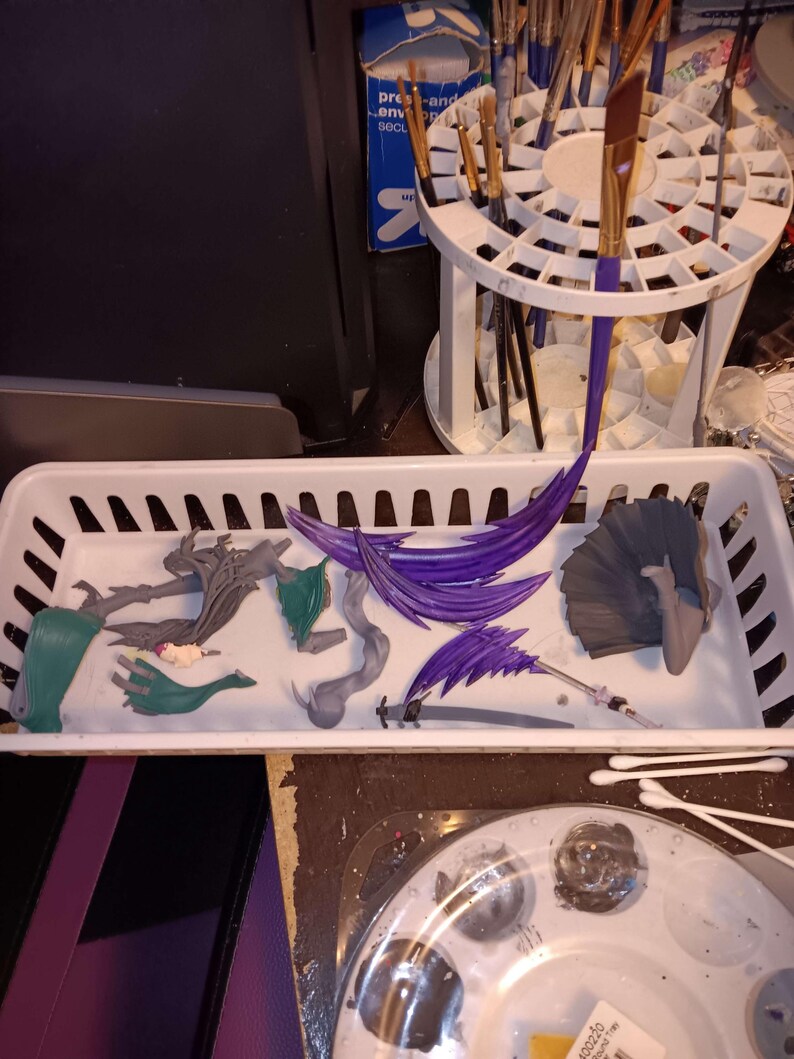 Puede incluir: Una bandeja de pl&aacute;stico blanco contiene varias figuras desmontadas y material de pintura. Las figuras son en tonos verdes, grises y morados. Los pinceles est&aacute;n en un soporte blanco. Una paleta de pintura con pintura gris est&aacute; en primer plano.