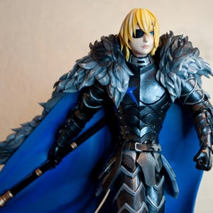 May include: A detailed figurine of a character in dark armour with a blue cape and blonde hair. The figure has a black eyepatch and is holding a staff. The armour is intricately detailed with silver and black accents, and the cape is a vibrant blue.