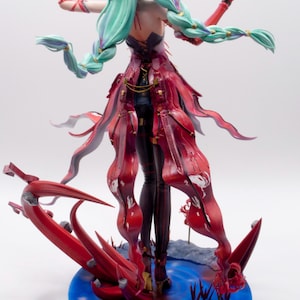 Puede incluir: Figura detallada de un personaje con largo cabello verde menta en trenzas, vistiendo un vestido rojo y negro. La figura posa sobre una base azul con elementos florales rojos estilizados. El personaje sostiene una flor roja.