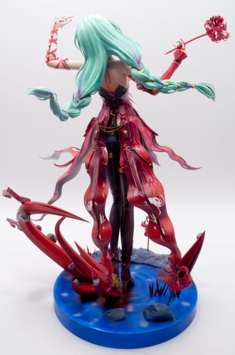 Puede incluir: Figura detallada de un personaje con largo cabello verde menta en trenzas, vistiendo un vestido rojo y negro. La figura posa sobre una base azul con elementos florales rojos estilizados. El personaje sostiene una flor roja.