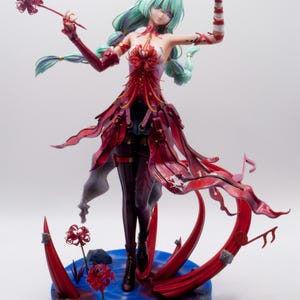 Puede incluir: Figura detallada de una mujer con cabello verde menta y un vestido rojo y negro. La figura est&aacute; posada sobre una base azul con detalles florales rojos. La figura es un objeto de colecci&oacute;n.