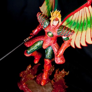 Op de afbeelding: Een rood en groen action figure met vleugels, die een zwaard vasthoudt, staand op een vurige basis. De figuur draagt ​​een harnas en een helm met een gouden kam.