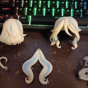 Puede incluir: Varias piezas de cabello blanco y azul claro de estilo anime están dispuestas sobre una superficie oscura. Las piezas incluyen dos secciones tipo peluca y un par de formas curvas en forma de ala. Un teclado es visible en el fondo.