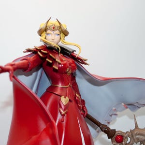 May include: A detailed figurine of a woman in a red dress with a flowing cape. The figure has blonde hair, a golden crown, and horns. She holds a staff with a red gem and a spiked head. The cape is light blue on the inside.