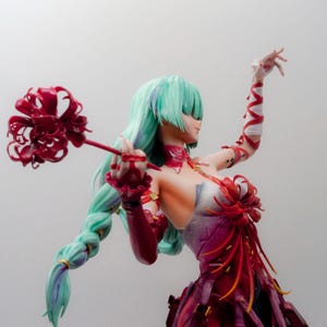 Puede incluir: Figura de una mujer con pelo largo verde claro en una trenza, vestida con un vestido rojo y morado. Sostiene una flor roja en una mano y tiene el otro brazo levantado. La figura tiene un dise&ntilde;o detallado y est&aacute; sobre un fondo liso.