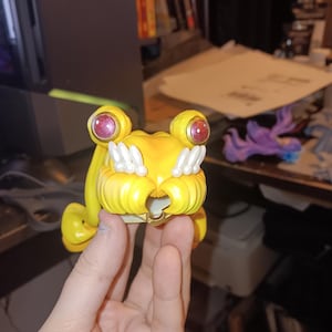 May include: A yellow figurine with large, round eyes and a detailed, textured design. The figurine has a stylized appearance with a bright yellow colour and accents of white and red. The overall design suggests a character or collectible item.