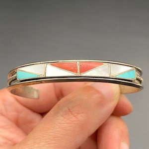 Op de afbeelding: Zilveren manchetarmband met ingelegde geometrische ontwerpen. De armband heeft turquoise, witte en koraalkleurige driehoekige inlegwerk. De metalen band heeft een gedraaid touwontwerp op de randen. Een klassiek sieraad.