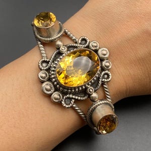 May include: A silver cuff bracelet featuring a large oval faceted yellow citrine gemstone at the center, flanked by two smaller round citrine stones. The metalwork includes twisted rope detailing and small silver spheres surrounding the stones.
