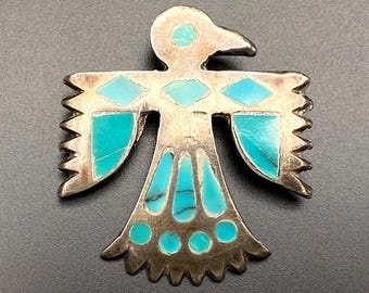 Puede incluir: Un broche de metal plateado con forma de ave estilizada con las alas extendidas. La pieza presenta incrustaciones de piedra color turquesa en formas geométricas, incluyendo diamantes, triángulos y círculos, decorando la cabeza, las alas y la cola.