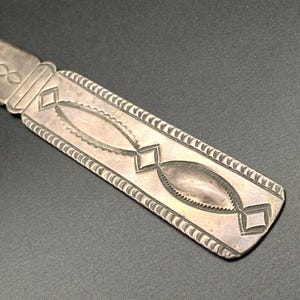 May include: Close-up of a silver metal object with detailed geometric patterns. The rectangular piece has diamond and oval shapes, with a decorative border. The metal has a slightly aged look, possibly vintage or antique.