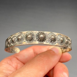 May include: Silver-toned cuff bracelet with a repeating pattern of stylized sunburst designs. The bracelet has an open design and is held by a hand against a gray background. The metal has a slightly aged appearance.
