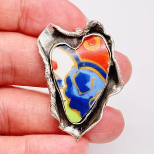 May include: A heart-shaped pendant featuring a colorful ceramic shard with blue, orange, yellow, and white patterns, encased in a textured, irregular silver-toned metal frame with a rough, organic edge and a metallic finish.