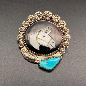 May include: A silver-toned brooch with a horse head design. The horse head is white against a black background, surrounded by a floral border. A turquoise stone is set at the bottom. The brooch has a leaf detail.