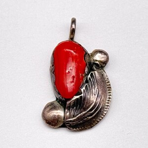 May include: A silver pendant featuring a smooth, vibrant red oval stone set within a textured metal frame. The design includes a small circular silver accent at the base and a curved, ridged silver element on the side, topped with a small bail.