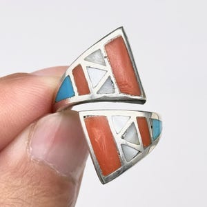 May include: A silver bypass ring featuring two triangular sections inlaid with geometric pieces of orange coral, white shell, and blue turquoise. The metal frame creates a segmented mosaic pattern across both sides of the open-ended ring design.