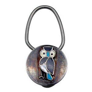May include: A silver-toned keychain with a circular pendant featuring an owl design. The owl has a black and gray body, white eyes, and turquoise accents. The keychain has a loop for attaching keys.