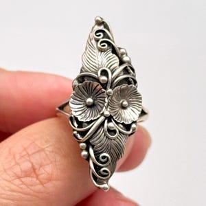 Könnte beinhalten: Ein silberner Ring mit floralem und Blattdesign. Der Ring hat zwei blütenförmige Elemente und detaillierte Blattakzente. Der Ring wird von einem Finger vor weißem Hintergrund gehalten.