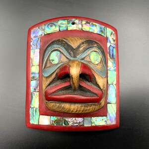 Peut inclure: Un masque en bois sculpté avec un cadre rouge et un motif de visage central. Le visage présente un nez proéminent, des lèvres rouges et des accents bleus et noirs. Le cadre est orné de morceaux de coquille d'ormeau irisés.