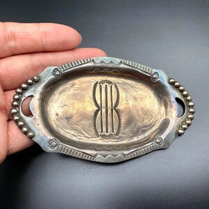 Puede incluir: Una bandeja ovalada de metal plateado con un diseño en relieve. La bandeja tiene un borde con cuentas y patrones decorativos a lo largo del borde. El centro presenta un diseño vertical estilizado. La bandeja es sostenida por una mano.