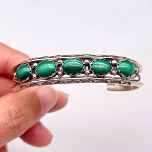 May include: A silver cuff bracelet with six oval, bright green malachite stones. The bracelet has an engraved design along the edges and around each stone, giving it a vintage or artisan appearance. The bracelet is held by a hand.
