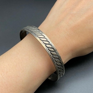 May include: A silver-toned metal cuff bracelet featuring a continuous twisted rope pattern along the centre. The band has a smooth, polished finish on the edges and a slightly oxidised appearance within the recessed areas of the rope design.
