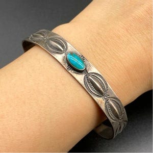 May include: A silver cuff bracelet featuring an oval turquoise stone. The bracelet has an engraved design with oval and floral patterns. The bracelet is on a wrist.